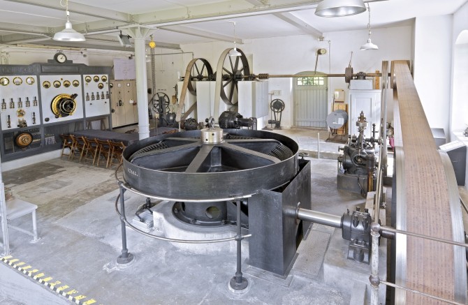 Hintergrund von links nach rechts: Jugendstil Schaltwand aus Marmor, Königswelle, Generator, Regulator. Vordergrund: Turbinenachse mit grossem Kegelzahnrad mit Holzzähnen